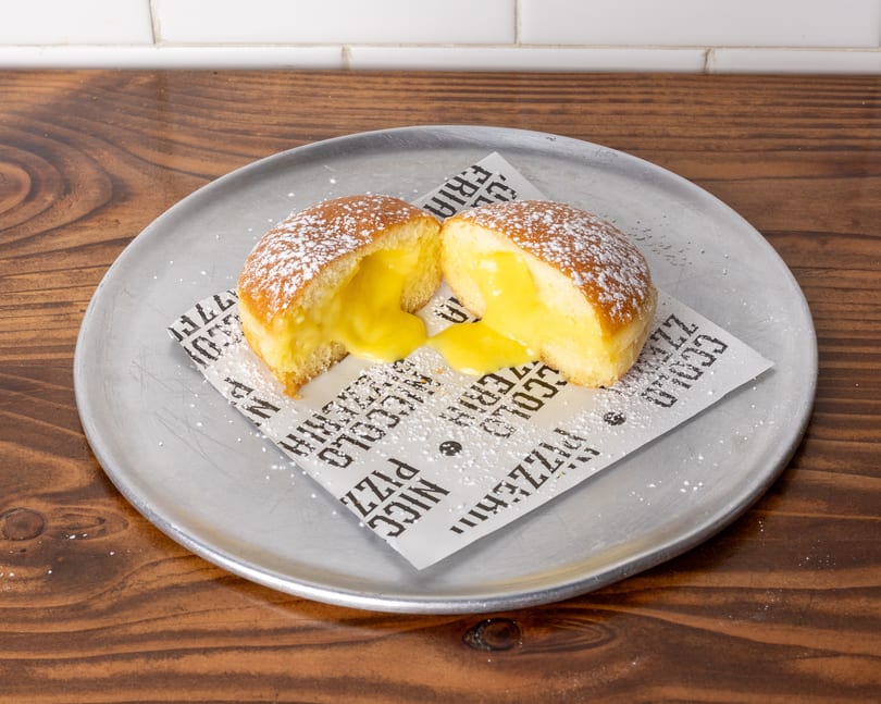 A powdered sugar-dusted pastry, split open to reveal a creamy yellow filling, on a metal plate.