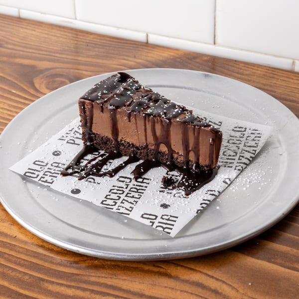 A slice of chocolate cheesecake with chocolate drizzle and powdered sugar on a metal plate.