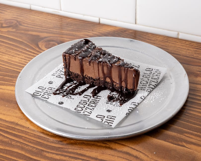 A slice of chocolate cheesecake with chocolate drizzle and powdered sugar on a metal plate.