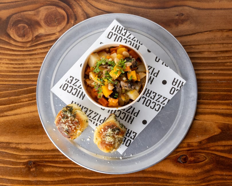 Hearty vegetable stew with white beans and herbs, served with two garlic knots on a metal tray.