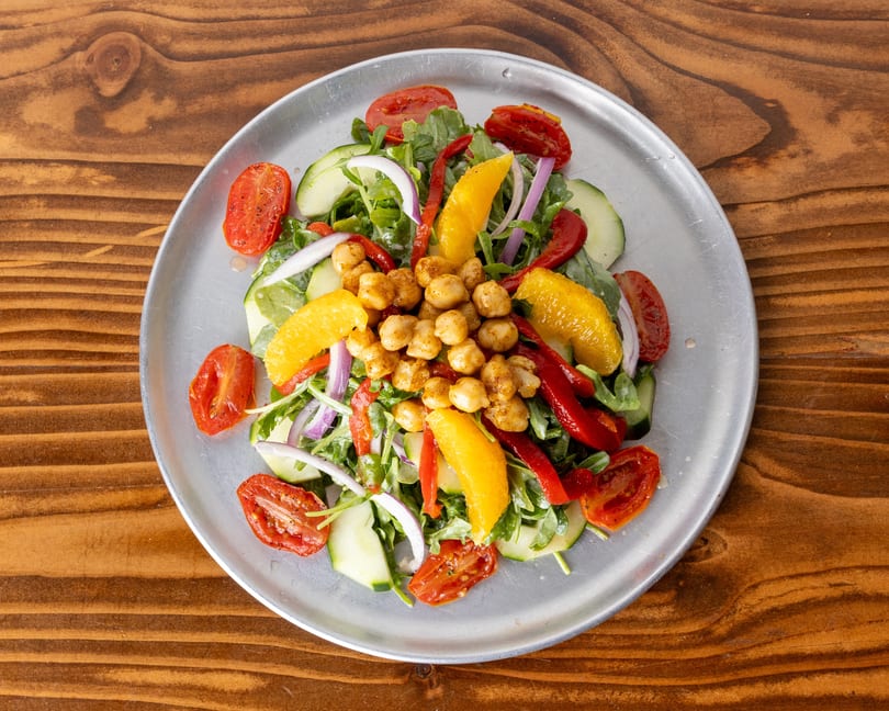 Colorful salad with chickpeas, roasted tomatoes, cucumbers, red onion, and orange segments on a metal plate.