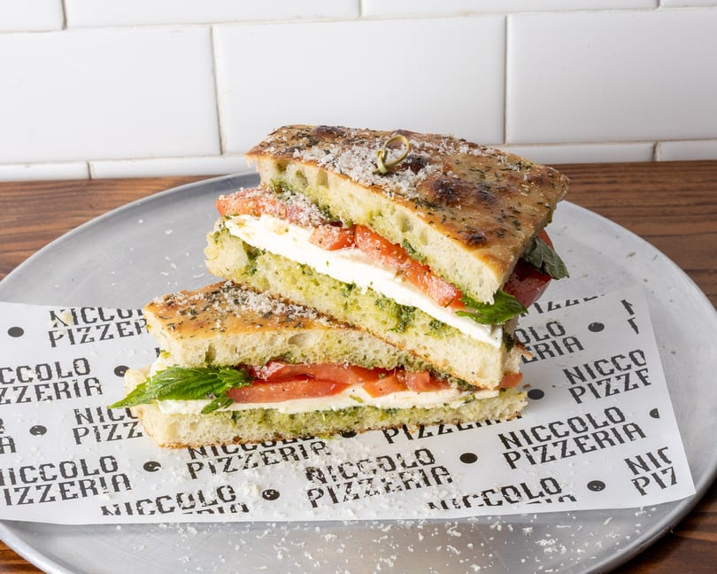 Two halves of a focaccia sandwich with pesto, mozzarella, and tomatoes on a metal tray.