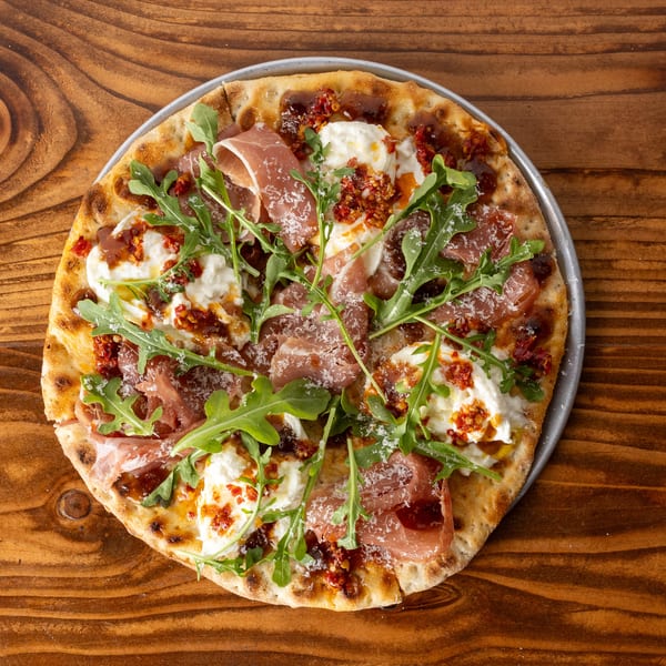 A round pizza with prosciutto, burrata cheese, arugula, and sun-dried tomatoes on a wooden table.