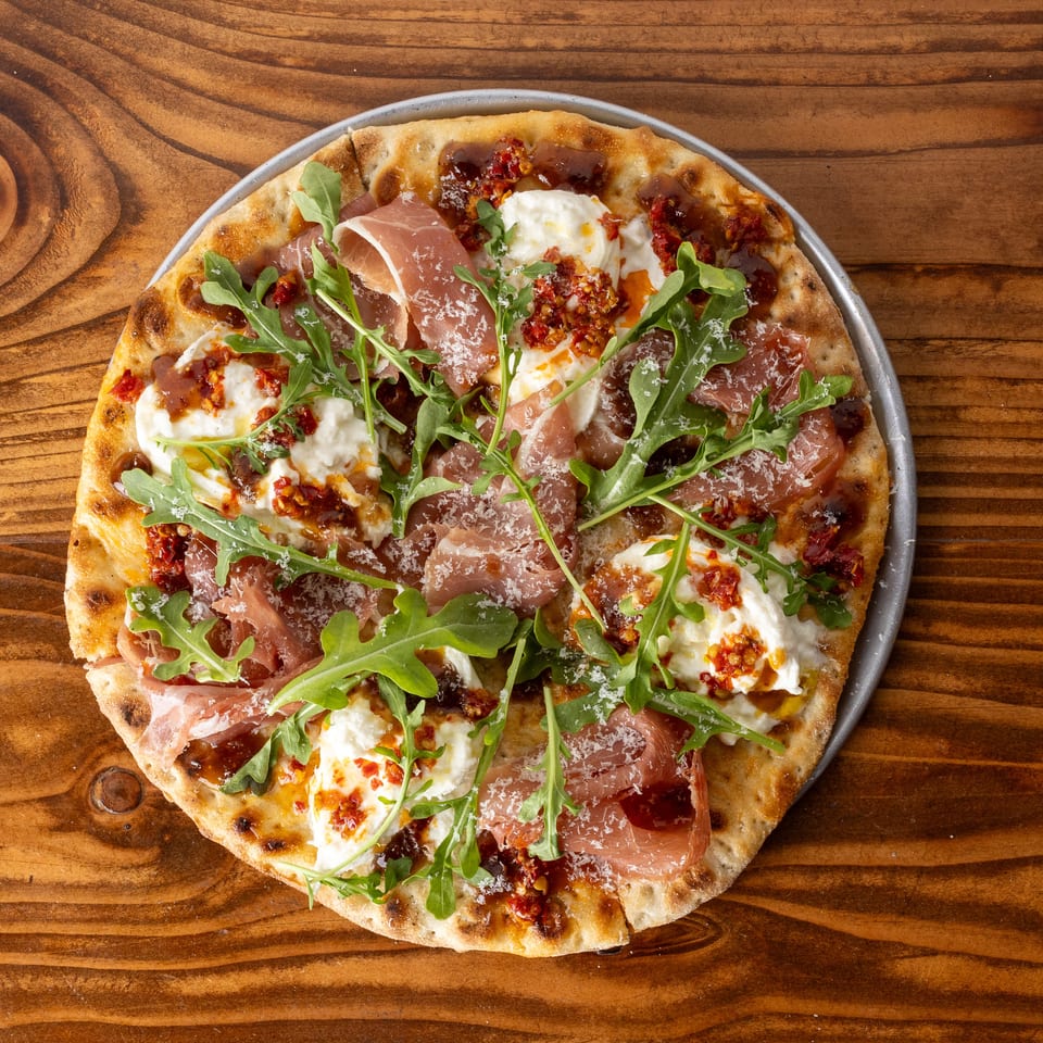 A round pizza with prosciutto, burrata cheese, arugula, and sun-dried tomatoes on a wooden table.