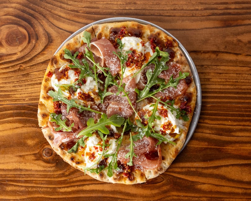 A round pizza with prosciutto, burrata cheese, arugula, and sun-dried tomatoes on a wooden table.