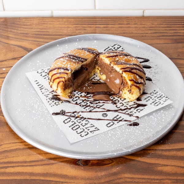 A split fried pastry filled with melted chocolate, drizzled with chocolate sauce and powdered sugar.
