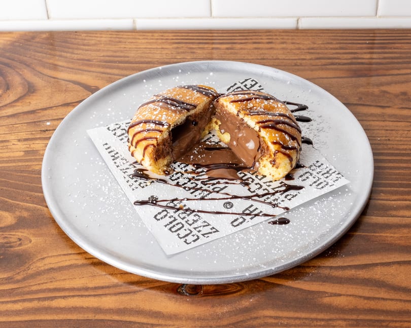 A split fried pastry filled with melted chocolate, drizzled with chocolate sauce and powdered sugar.