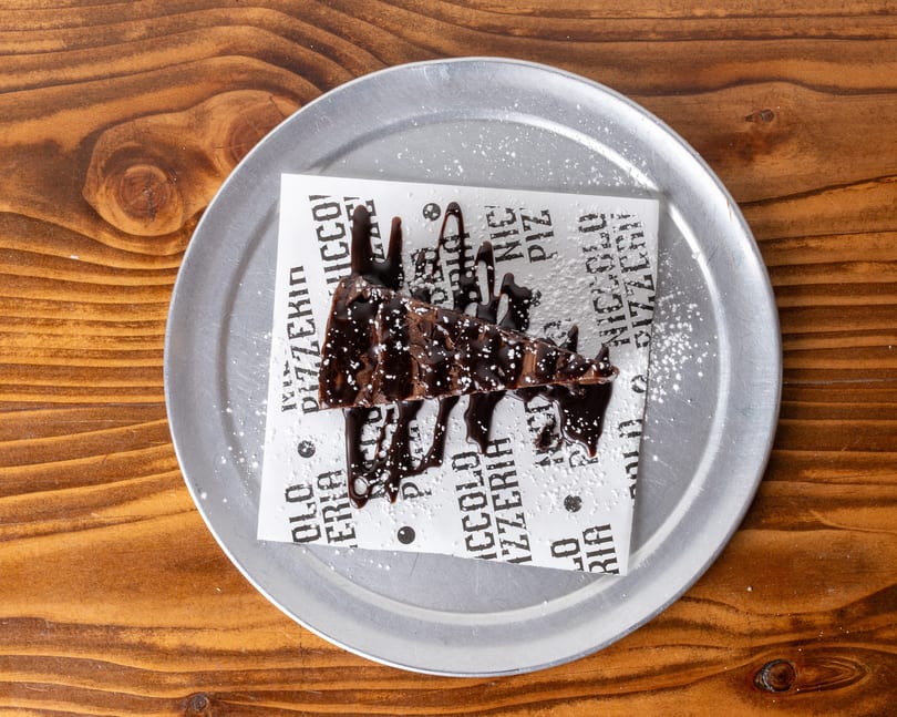 A chocolate brownie slice with chocolate drizzle and powdered sugar on a metal plate.