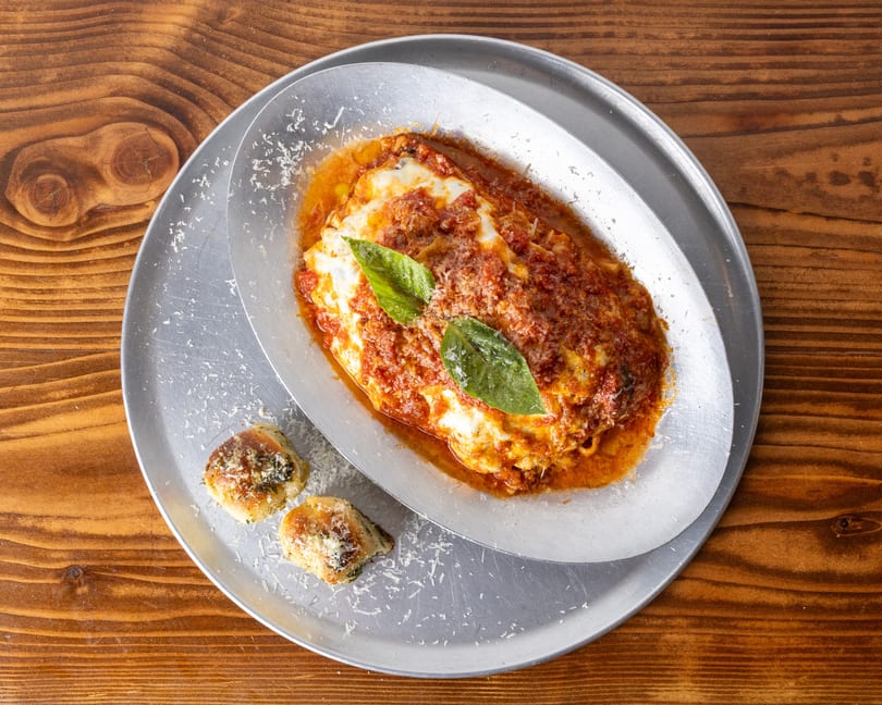 Baked lasagna with rich red sauce, melted cheese, fresh basil, and two garlic knots on a metal tray.