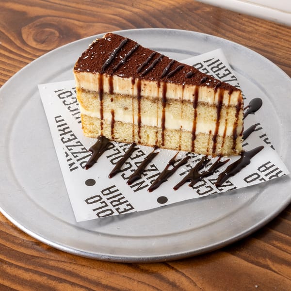 A slice of tiramisu with chocolate drizzle and powdered sugar on a metal plate.
