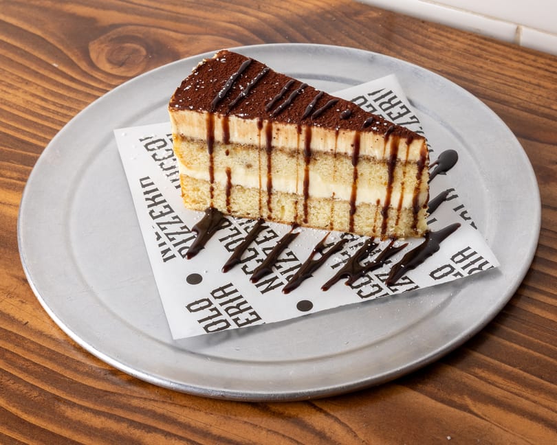 A slice of tiramisu with chocolate drizzle and powdered sugar on a metal plate.