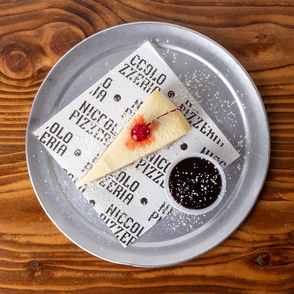Cheesecake slice with a cherry and chocolate sauce, dusted with powdered sugar on a metal tray.