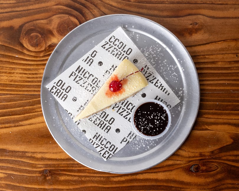 Cheesecake slice with a cherry and chocolate sauce, dusted with powdered sugar on a metal tray.